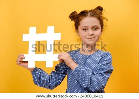 Portrait of funny adorable little girl child holding big white hashtag symbol, happy looking at camera, popular blogging, child content, posing isolated over plain yellow studio background wall Royalty-Free Stock Photo #2406010411