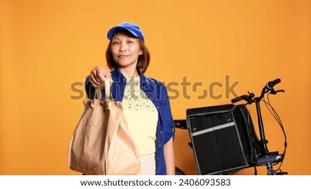 Slow motion portrait shot of friendly courier handing lunch bag to customer, showing thumbs up sign. Food delivery cyclist providing meal order to client, isolated over studio background