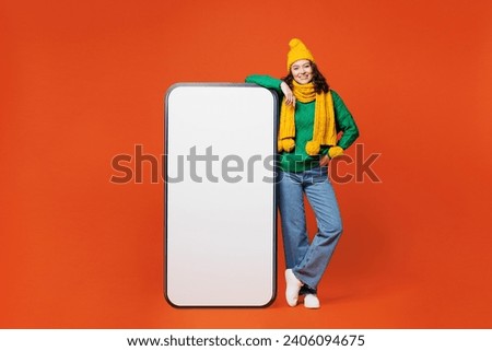 Full body young smiling cheerful woman she wear green knitted sweater yellow hat scarf stand near big huge blank screen mobile cell phone smartphone with area isolated on plain orange red background