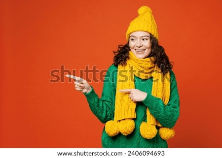 Young happy woman she wear green knitted sweater yellow hat scarf pointing index finger aside indicate on workspace area copy space isolated on plain orange red background studio. Lifestyle concept