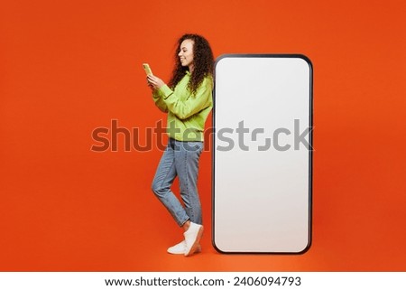 Full body sideways young woman of African American ethnicity wear green hoody casual clothes big huge blank screen mobile cell phone with area use smartphone isolated on plain red orange background
