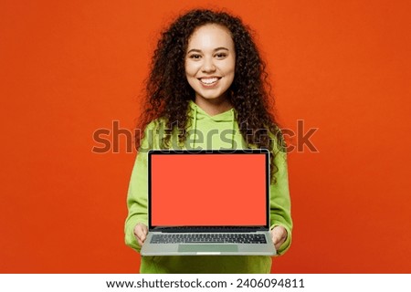 Smiling fun young IT woman of African American ethnicity she wear green hoody casual clothes hold use work on blank screen laptop pc computer isolated on plain red orange background. Lifestyle concept