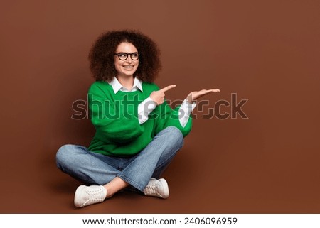 Full size photo of charming girl sit floor indicate finger hand hold empty space isolated on brown color background
