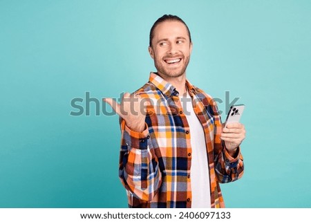 Photo of cheerful nice man dressed plaid shirt hold smartphone look directing at promo empty space isolated on teal color background