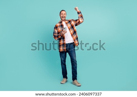 Full size photo of satisfied man dressed checkered shirt jeans hold smartphone raising fist win gamble isolated on teal color background