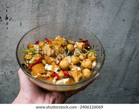 Close-up of a large bowl held by a person, containing food with pieces of tofu, small balls, and red and green chilies