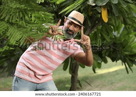 Portrait of cheerful adult man showing photo frame gesture at the park. Man wearing round hat outdoors in nature park make photo frame gesture.