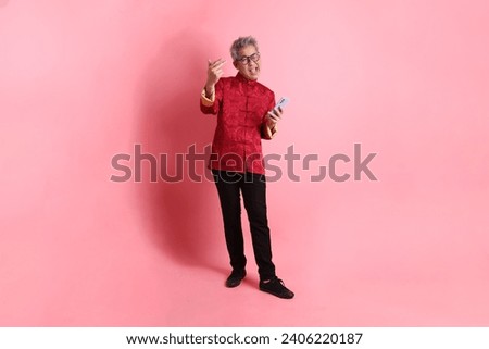 Happy Chinese new year. Asian Chinese energetic senior man wearing red traditional cheongsam qipao or changshan dress with hand holds mobile phone isolated on pink background.