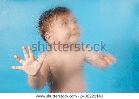 Baby behind glass, on blue background. Water drops on glass, Blurred image of child through foggy glass, concept.