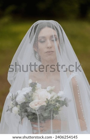 The bride in a lush dress and veil, holding a bouquet of white flowers and greenery, posing against the background of green trees.. Spring wedding