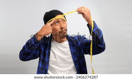 a curious Asian man, dressed in a beanie hat and casual shirt, uses a measuring tape to measure the diameter of his head while standing against white background. Royalty-Free Stock Photo #2406274287