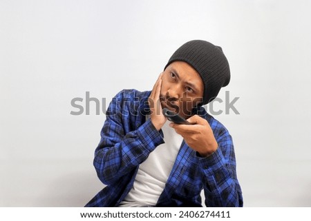 Bored young Asian man, dressed in a beanie hat and casual shirt, holds a TV remote control, flipping through different channels in search of an interesting program while watching TV, white background