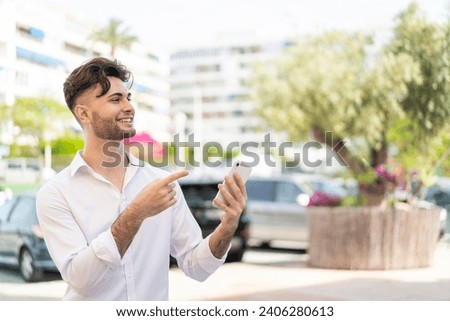 Young handsome man using mobile phone at outdoors pointing to the side to present a product