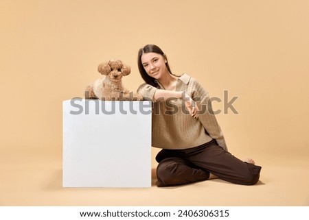 Full length portrait of young attractive lady sitting near white cube where sitting her purebred dog, poodle against beige background. Concept of animal, pet lover, friendship, domestic life. Royalty-Free Stock Photo #2406306315