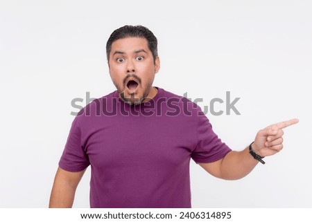 A middle aged man looks flabbergasted, looking at the camera while pointing to the right. Wearing a purple waffle shirt. Isolated on a white background. Royalty-Free Stock Photo #2406314895