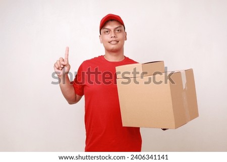 Happy Asian courier man in red holding a cardboard box while showing a finger. Isolated on white