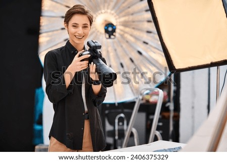 cheerful attractive woman in casual attire smiling at camera and holding camera in her photo studio