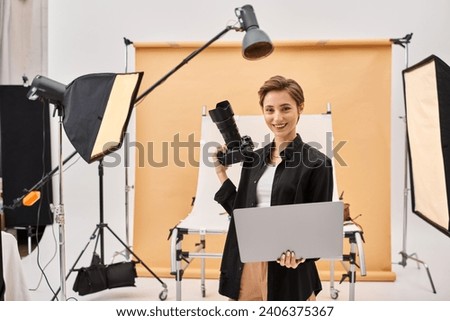 cheerful attractive woman smiling happily while retouching photos and holding her camera in studio
