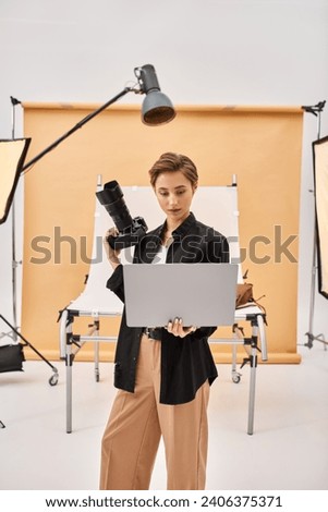good looking young woman in casual attire retouching photos using her laptop and holding camera