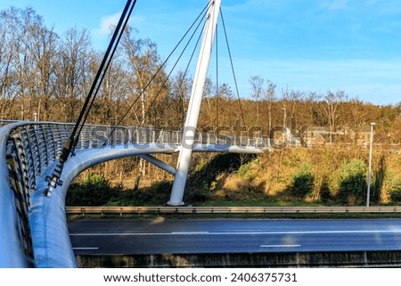 Part of cable-stayed bridge for bicycles and pedestrians over highway towards Hoge Kempen national park, bare trees against blue sky in background sunny autumn day in As Limburg, Belgium Royalty-Free Stock Photo #2406375731