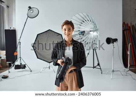happy pretty woman with short hair in everyday clothes posing in her studio with camera in hands Royalty-Free Stock Photo #2406376005