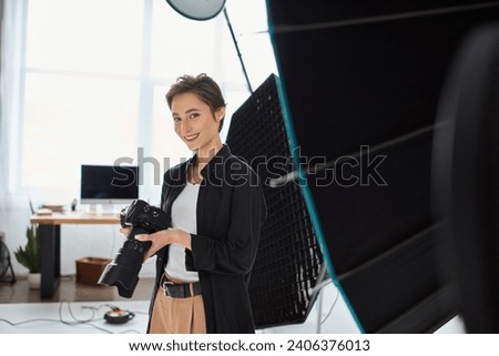 jolly pretty woman with short hair in everyday clothes posing in her studio with camera in hands