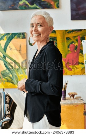 stylish and cheerful middle aged woman artist looking at camera near creative paintings in studio Royalty-Free Stock Photo #2406378883
