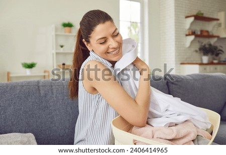 Young housewife smells clean clothes. Happy, smiling woman sitting on the couch with a laundry basket, holding a clean, fresh, white shirt and smelling a nice, pleasant smell of the fabric conditioner Royalty-Free Stock Photo #2406414965