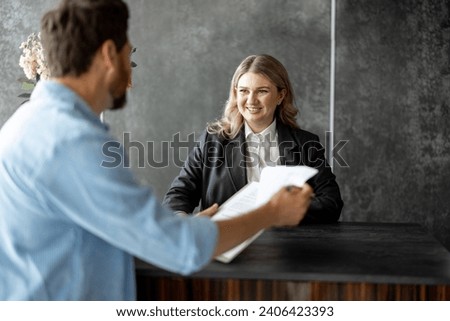Young medical receptionist talks with a male client and gives some paper to sign at the reception desk of modern clinic