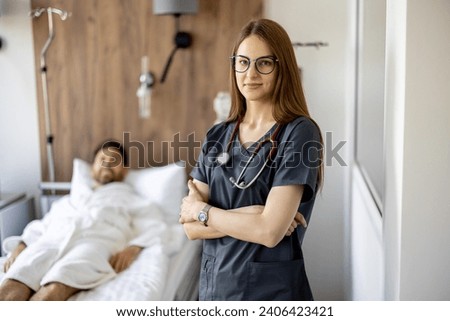 Portrait of a young female nurse standing in medical ward with patient resting on background. Nursing and medical support concept