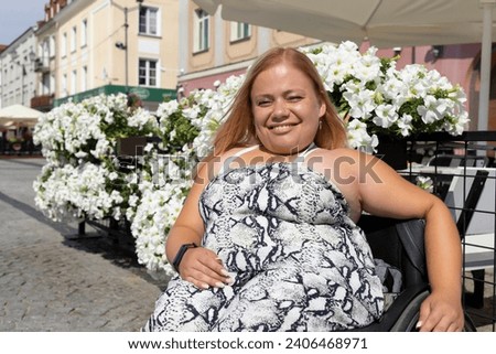 Smiling Brightly Young Woman With Short Stature Sitting In Wheel Chair In Street Outdoor. Flowers, Cafe On Background. Person With Special Needs Enjoys Social Life. Horizontal. High quality photo Royalty-Free Stock Photo #2406468971