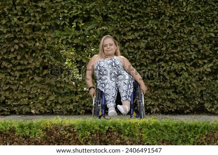 International Wheelchair Day , March 1st Smiling. Pretty Young Woman With Short Stature Sitting In Wheel Chair In Outdoor. Green Wall On Background. Person With Special Needs Enjoys Social Life. Royalty-Free Stock Photo #2406491547