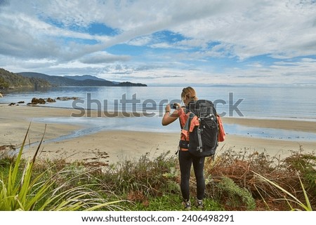 Woman hiking with backpack arriving to sandy beach in New Zealand, Stewart Island, North West Circuit. Taking a picture with cell phone camera