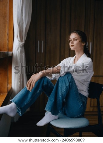 Woman sitting at home by a wooden window with a smile on a chair in homemade comfortable clothes and looking at the landscape, spring mood, women's day, rest on the weekend. Royalty-Free Stock Photo #2406598809