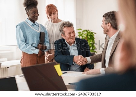 Multi-ethnic business professionals shaking hands in agreement, with attentive colleagues in the background. Royalty-Free Stock Photo #2406703319