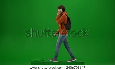 Portrait of person tourist isolated on chroma key green screen background. Young man in orange hoodie with backpack walking and talking on smartphone.