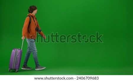 Portrait of person tourist isolated on chroma key green screen background. Young man with suitcase walking with suitcase in orange hoodie and jeans.