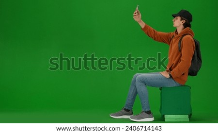 Portrait of person tourist isolated on chroma key green screen background. Young man sitting holding smartphone and taking selfie pictures.