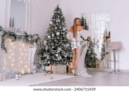 The blonde in the Christmas room. A beautiful blonde woman in a shiny light short dress with a train stands in a beautiful bright room decorated with a festive interior with a Christmas tree.