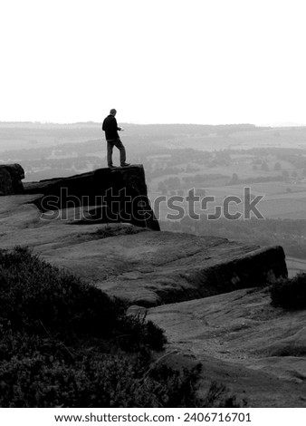 A man standing on the jagged edge of the cliff overlooking the landscape, and studying a map.  Curbar Edge, Peak District, Derbyshire, UK.  Countryside navigation; planning his route.  September 2009. Royalty-Free Stock Photo #2406716701