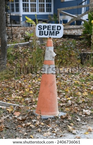 An old orange and white traffic cone set off the side of the road that has a small black and white sign on top of it that says "Speed Bump".