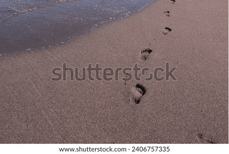 Foot prints in black sand on the island of Hawaii along the shoreline with the waves lapping from the Pacific Ocean from several angles.  Blue skies on a sunny day in this beautiful tropical location.