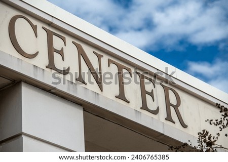 A diagonal sign with the word centre made of brown brass letters on an exterior wall of a contemporary commercial style building with aluminum metal composite panels. Sun and clouds in the back.