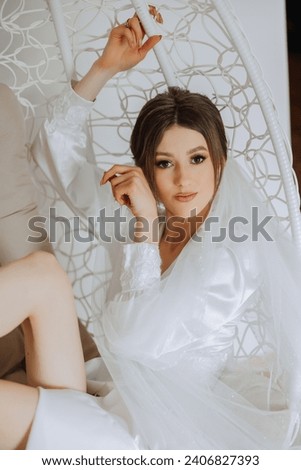 Morning of the bride in boudoir style. A woman bride in a white robe sits on a wicker swing near her wedding dress on a mannequin. Wedding day. Royalty-Free Stock Photo #2406827393