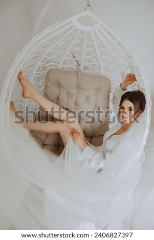 Morning of the bride in boudoir style. A woman bride in a white robe sits on a wicker swing near her wedding dress on a mannequin. Wedding day.