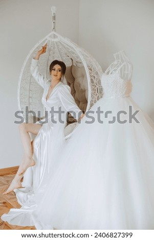 Morning of the bride in boudoir style. A woman bride in a white robe sits on a wicker swing near her wedding dress on a mannequin. Wedding day. Royalty-Free Stock Photo #2406827399