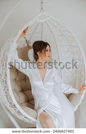 Morning of the bride in boudoir style. A woman bride in a white robe sits on a wicker swing near her wedding dress on a mannequin. Wedding day. Royalty-Free Stock Photo #2406827401