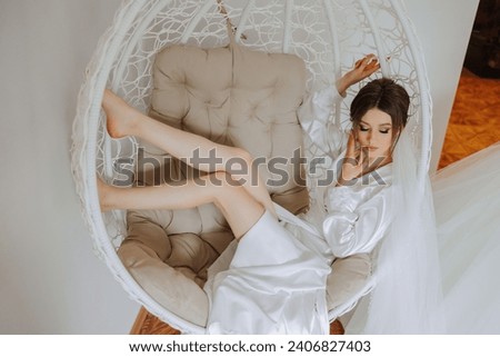 Morning of the bride in boudoir style. A woman bride in a white robe sits on a wicker swing near her wedding dress on a mannequin. Wedding day. Royalty-Free Stock Photo #2406827403