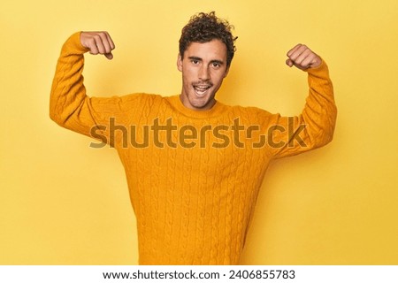 Young Latino man posing on yellow background showing strength gesture with arms, symbol of feminine power
