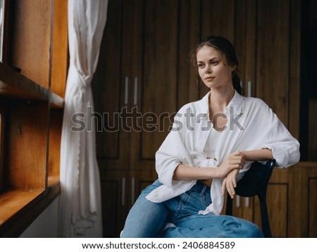 Woman sitting at home by a wooden window with a smile on a chair in homemade comfortable clothes and looking at the landscape, spring mood, women's day, rest on the weekend. Royalty-Free Stock Photo #2406884589